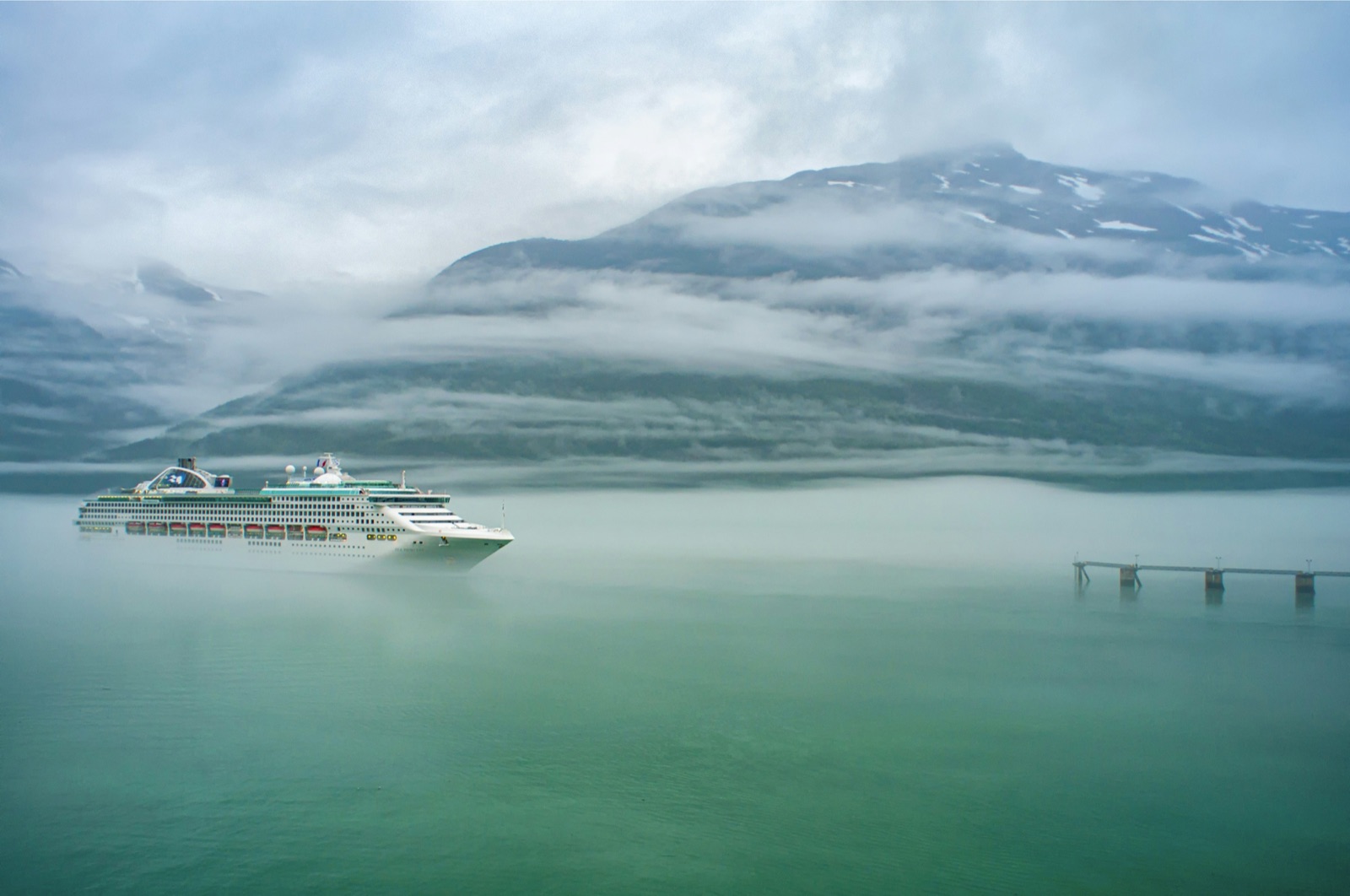 A white cruise ship in daylight on the water off Skagway, low cloud hanging over the mountains