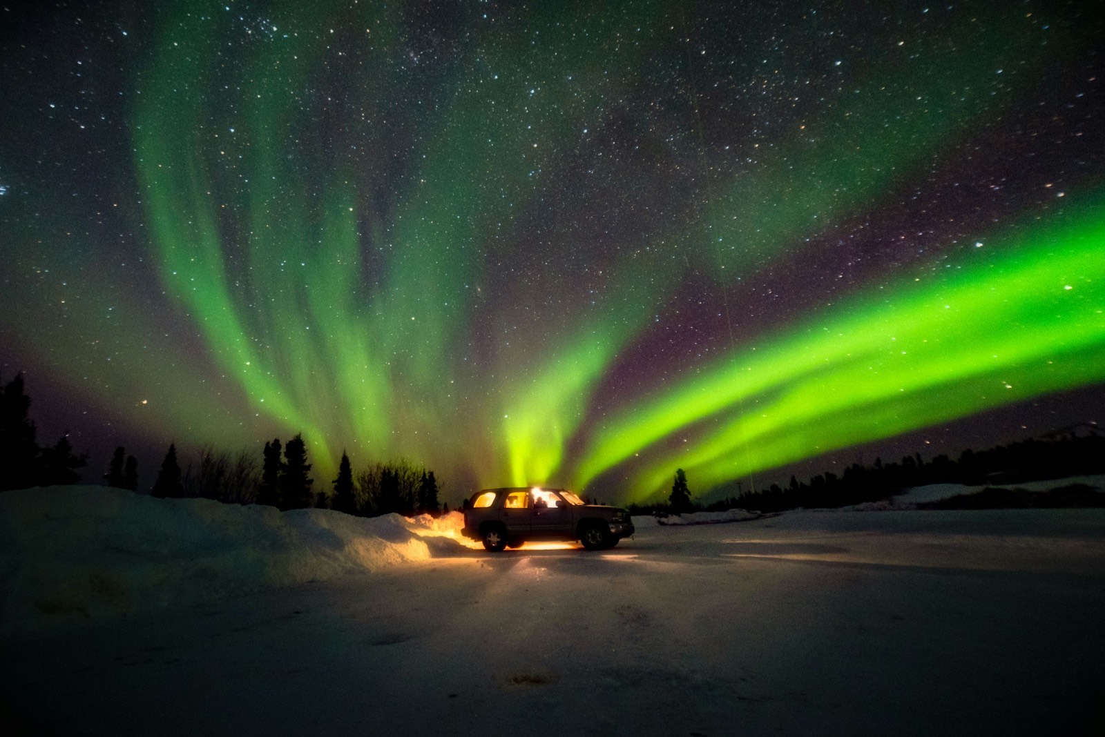 A car with its headlights on in the snow under bright green and purple aurora near Fairbanks