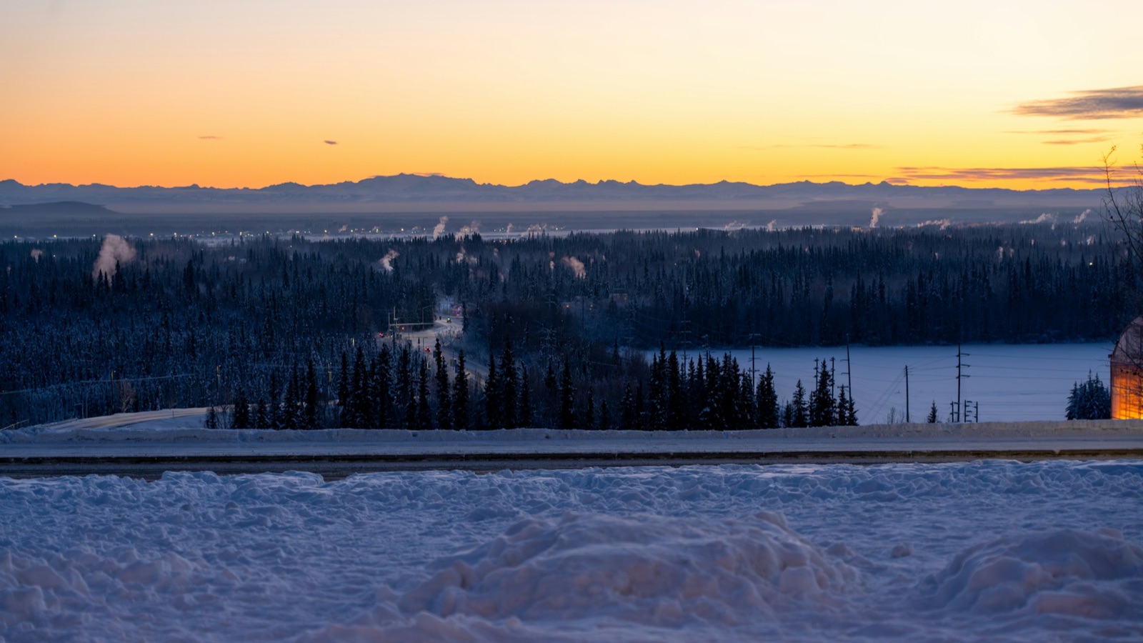 Orange winter twilight over snowy spruce forest in Fairbanks, with steam plumes rising from town and mountains on the horizon