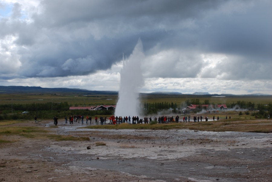 Strokkur geyser shooting a column of water into a stormy sky as a crowd watches