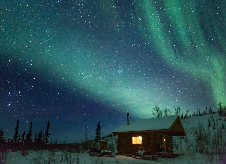 A lit log cabin on a snowy hillside under a wide green aurora and a starry sky