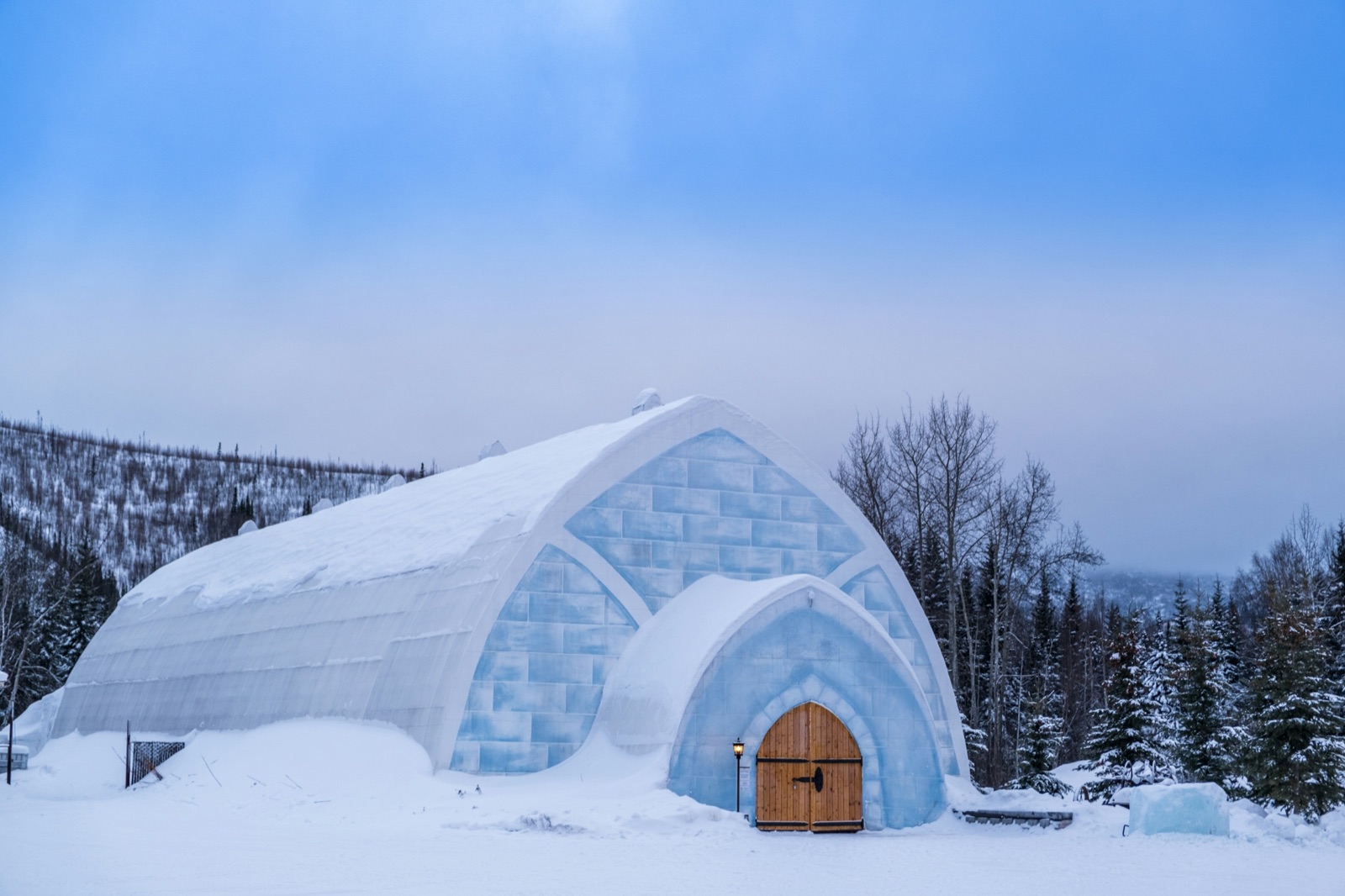 The arched, ice-block entrance of the Aurora Ice Museum building, with a wooden door, in snow under a blue winter sky