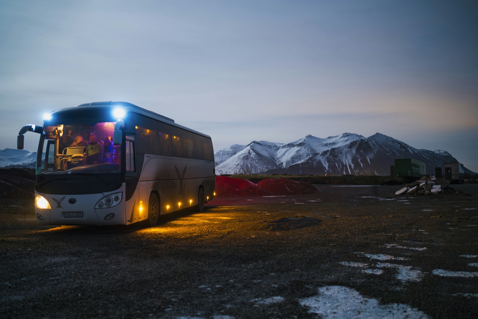 A northern lights tour coach parked with its lights on at dusk, snowy mountains behind