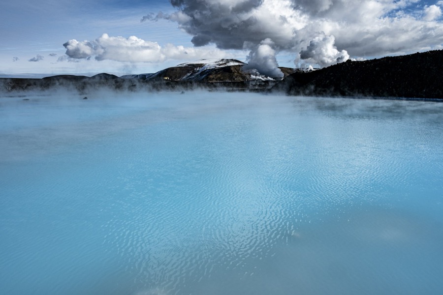Steam drifting across the milky blue water of the Blue Lagoon, with a black lava field and the plumes of the geothermal plant behind