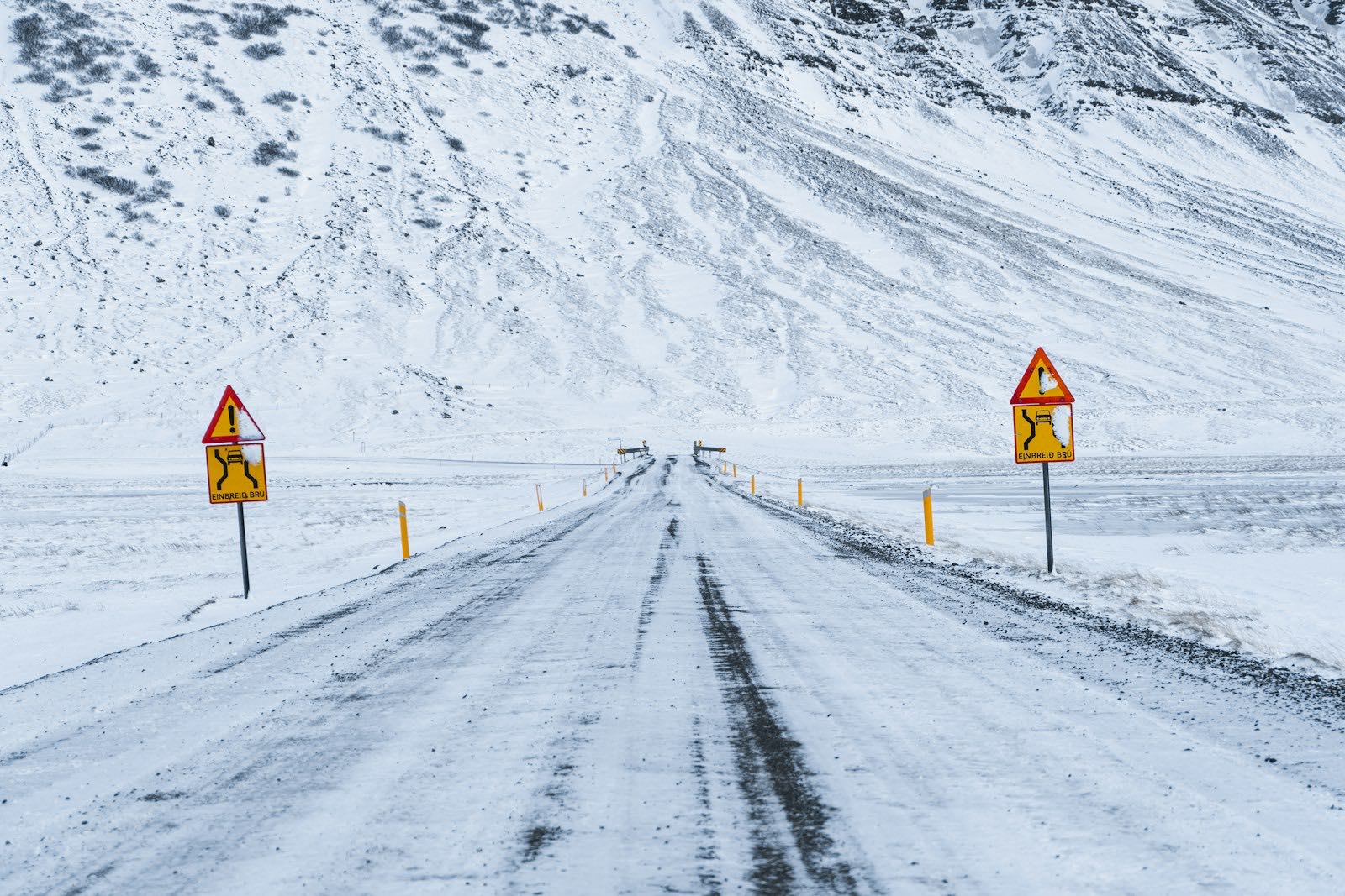 A snow-packed road in Iceland running to a single-lane bridge, with yellow 'Einbreið brú' warning signs on both sides and a snowy mountainside behind