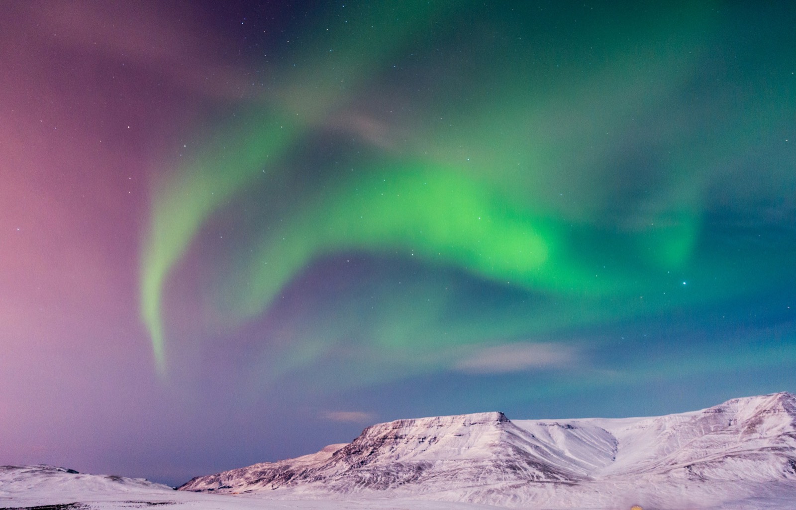 Swirls of green and pink aurora over a snow-covered mountain near Reykjavík