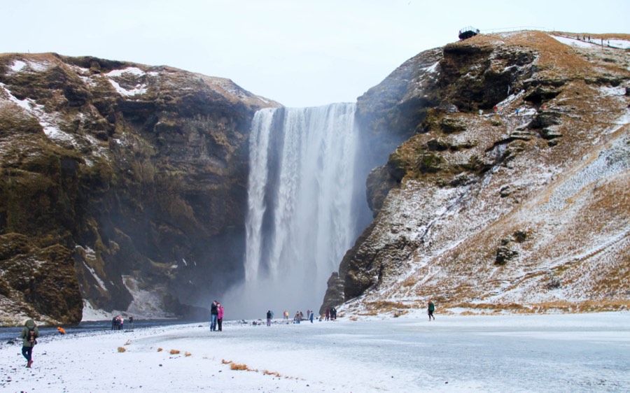 Skógafoss waterfall pouring between snow-streaked cliffs, with visitors on the frozen ground below